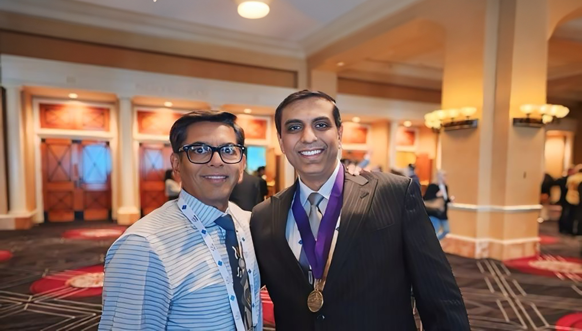 The image shows two individuals posing together for a photograph, with one person wearing a medal around their neck and the other standing beside them. They are indoors, possibly in a conference or event setting, as indicated by the background which features a stage with lighting equipment and a red carpet.